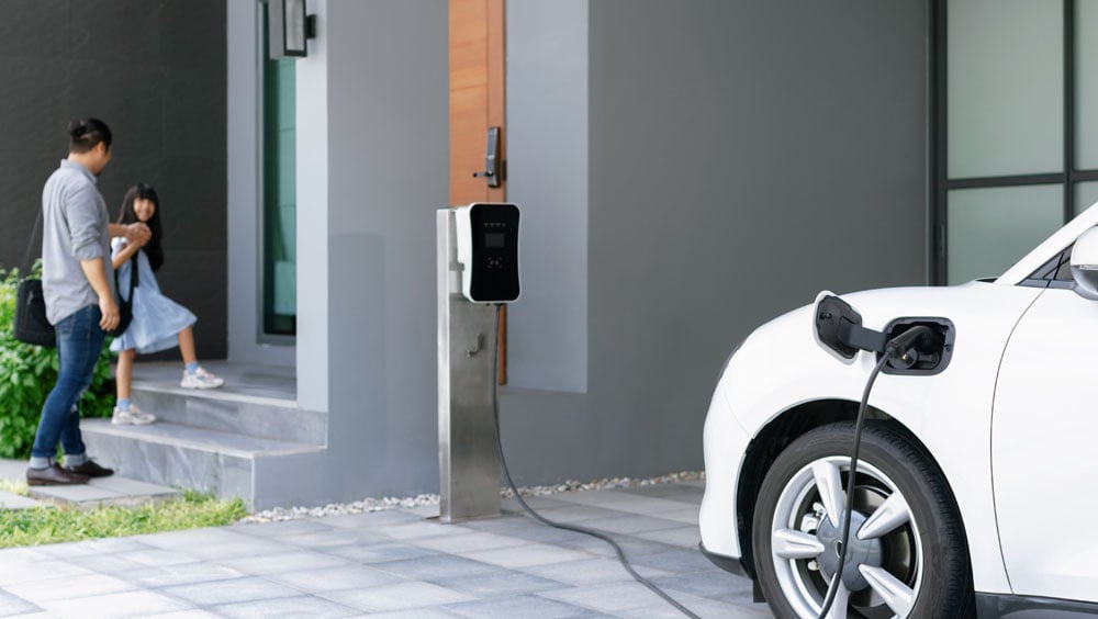 A white electric car charging at a wallbox in front of a residential building, in the background a man with a child - symbolizes data-based e-car advice and the consideration of individual employee needs