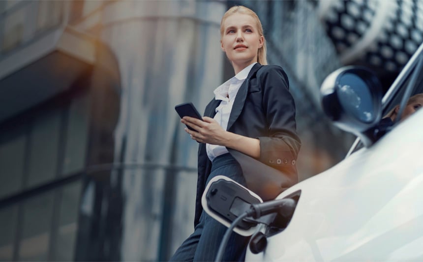 Businesswoman leaning relaxed against an electric car with charging cable in front of a modern office building - a symbol of e-mobility and innovation.