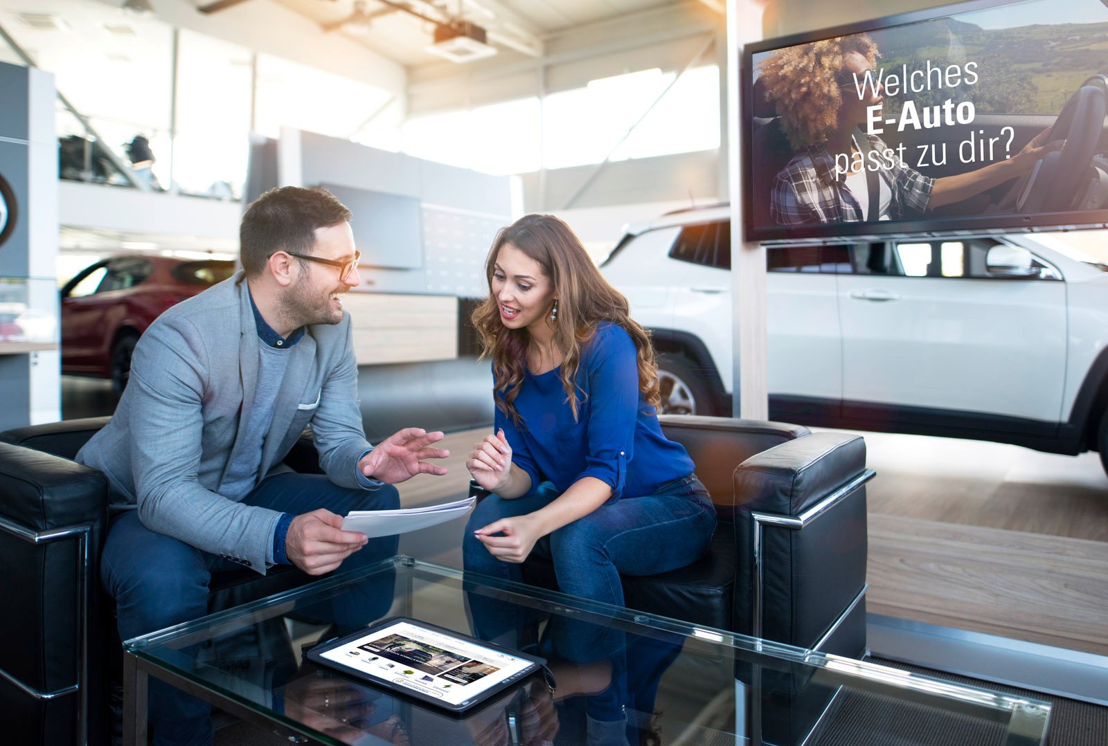 A man and a woman discussing a document, a symbol of holistic advice on electromobility; a tablet with digital solutions on the table, an electric car in the background