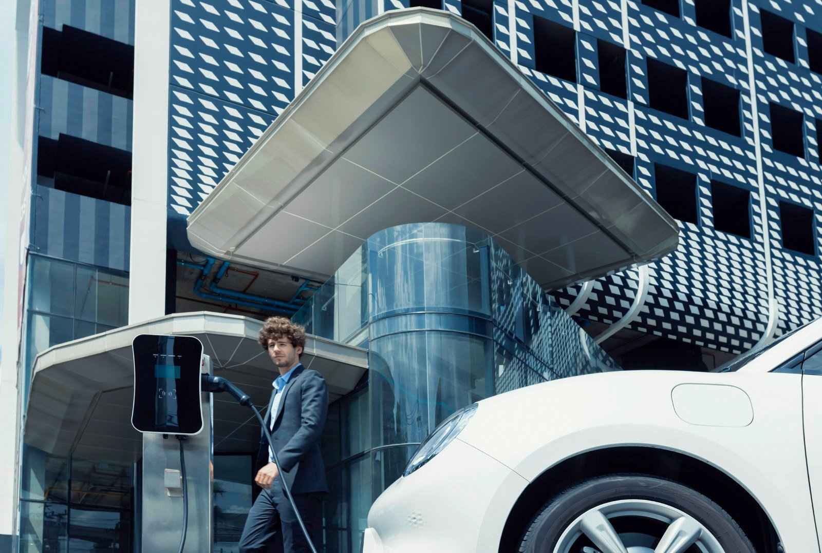 Man in a suit stands at a charging station next to a white electric car in front of a modern office building - symbol for e-Fleet solutions in commercial customer sales
