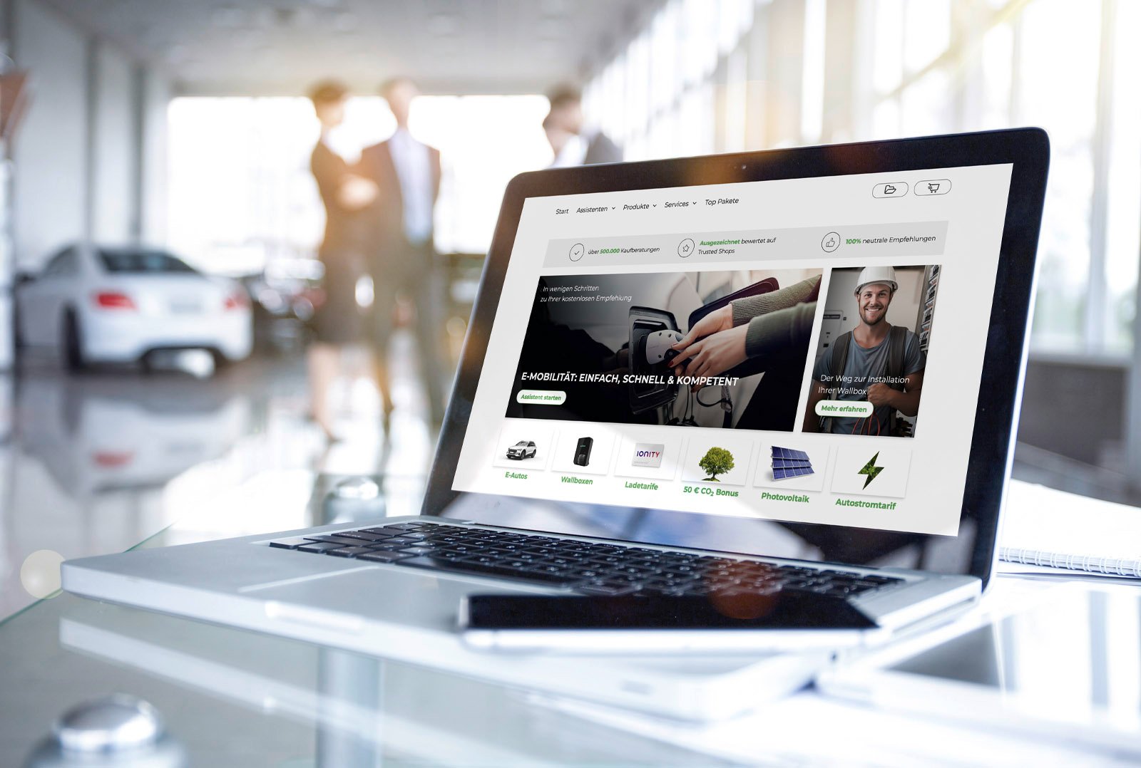 Laptop on glass table with open e-mobilio e-purchase advisor, in the background car dealership with blurred people - symbol for digital sales advice on electric mobility in private customer sales